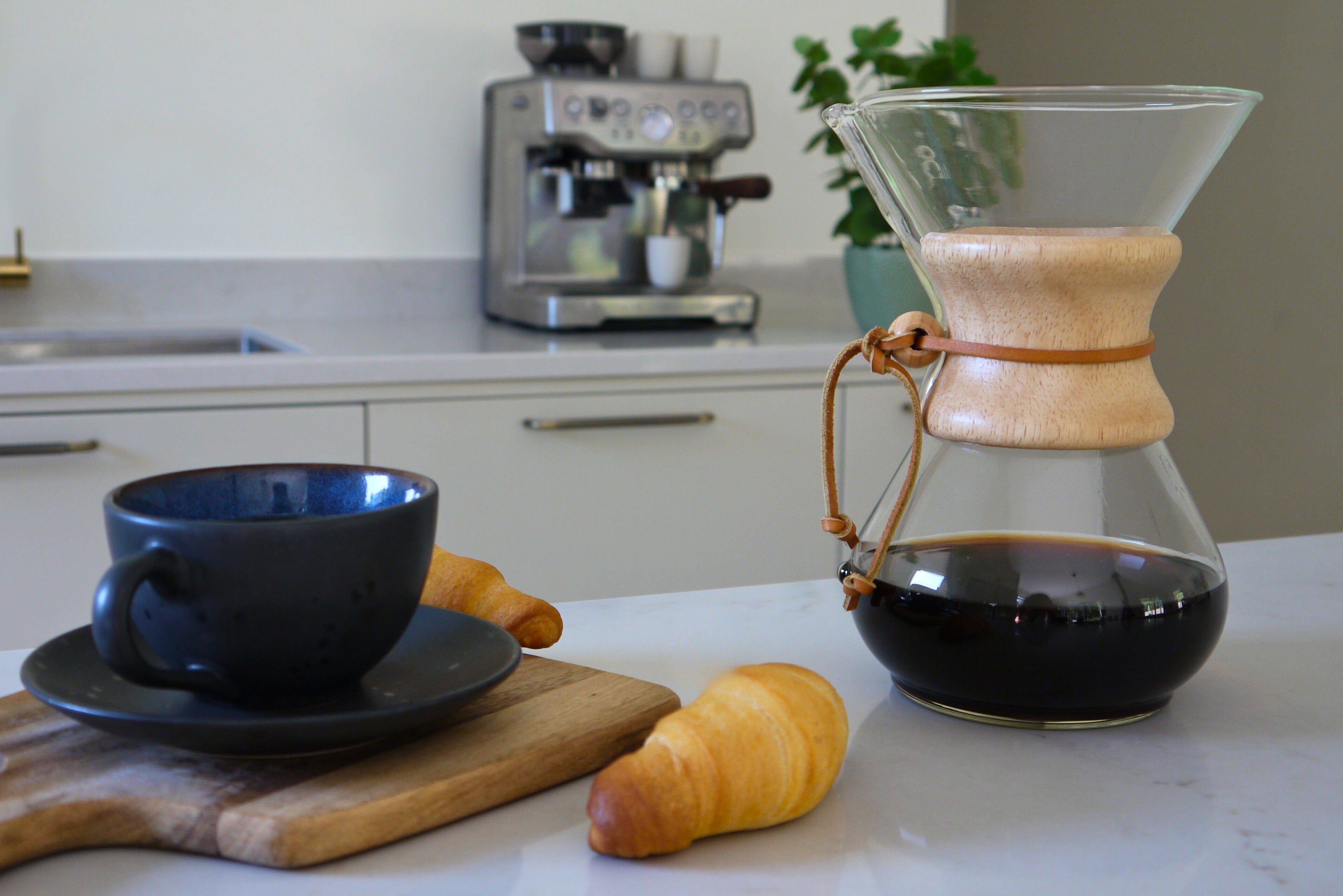 Coffee setup with a Chemex coffee maker, black cup, and croissant on a kitchen counter.