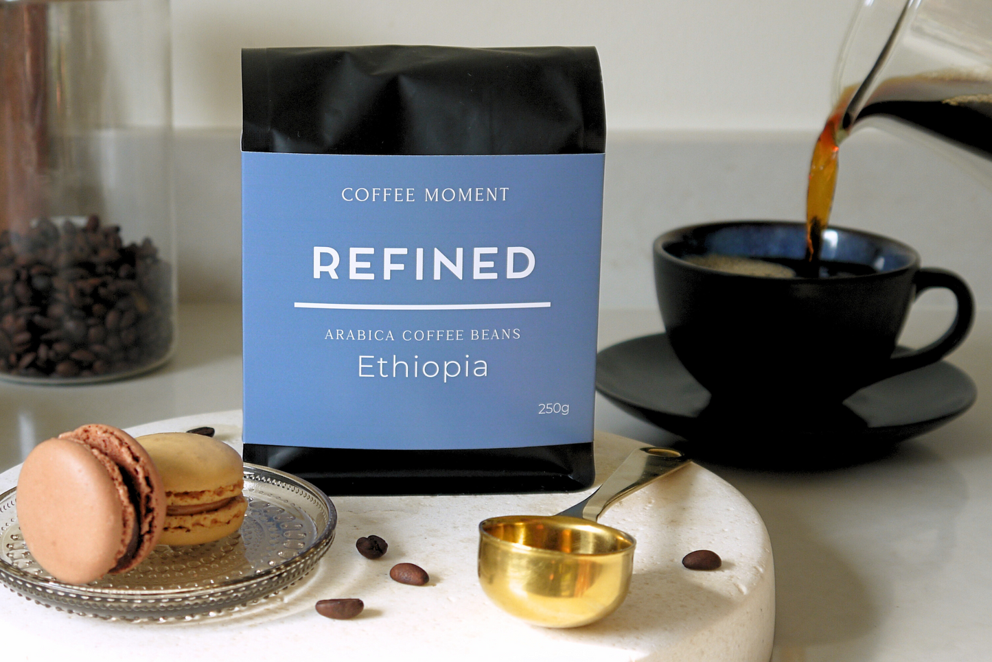 Coffee bag labeled 'Refined' with coffee beans, a macaron, and a cup on a table.